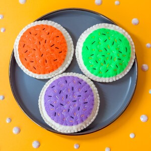 Op de afbeelding: Drie kleurrijke vilten koekjes op een grijze plaat tegen een gele achtergrond. De koekjes zijn oranje, groen en paars, elk met geborduurde hagelslag en een witte rand. Decoratieve elementen zijn verspreid.