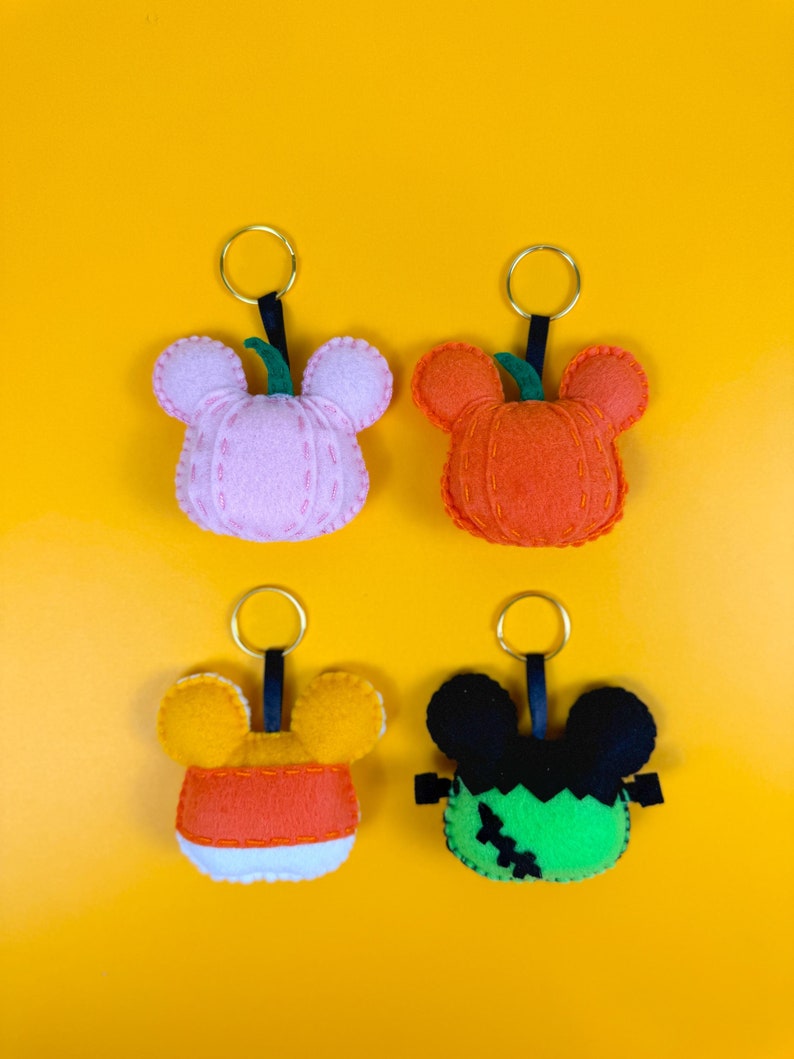 May include: Four felt Halloween keychains with Mickey Mouse ears. The keychains are shaped like a pumpkin, a candy corn, a Frankenstein monster, and a pink pumpkin.