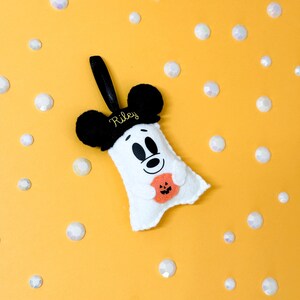 Op de afbeelding: Een wit vilt spookvormig ornament met zwarte Mickey Mouse oren en een zwart handvat. De geest heeft een lachend gezicht en houdt een oranje pompoen vast. De naam "Riley" is op de hoed geborduurd. Gele achtergrond met witte iriserende edelstenen.