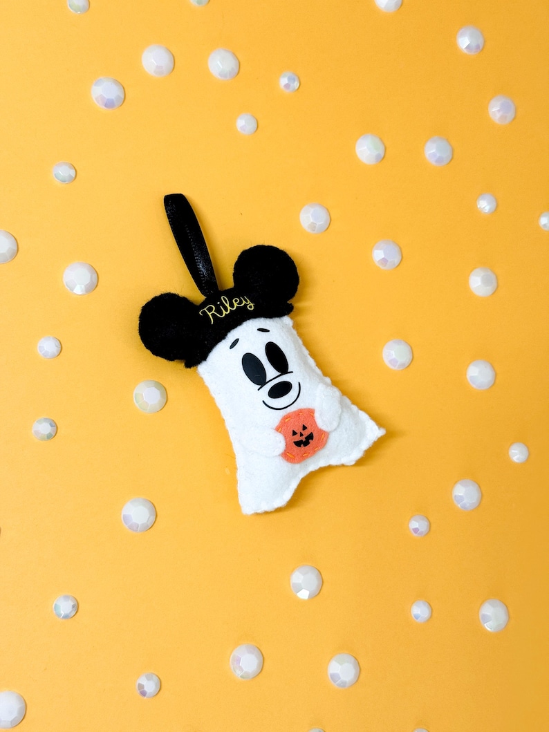 Op de afbeelding: Een wit vilt spookvormig ornament met zwarte Mickey Mouse oren en een zwart handvat. De geest heeft een lachend gezicht en houdt een oranje pompoen vast. De naam "Riley" is op de hoed geborduurd. Gele achtergrond met witte iriserende edelstenen.