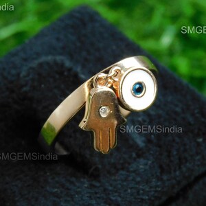 May include: Gold ring with a hamsa hand charm and a blue evil eye charm. The ring is on a black velvet display.