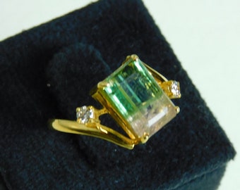 Può includere: Un anello d'oro con una grande pietra preziosa verde e bianca e due diamanti più piccoli. L'anello è esposto su una scatola di velluto nero.