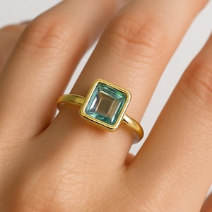 Op de afbeelding: Een gouden ring met een vierkante, lichtblauwe edelsteen. De edelsteen is gezet in een vierkante bezel en de band is van gepolijst goud. De ring wordt op een vinger getoond.