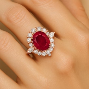 Puede incluir: Anillo de oro rosa con una gran piedra central de rubí ovalada, rodeada por un halo de diamantes redondos más pequeños. Acentos de diamantes adicionales están engastados en los lados, creando un diseño de racimo. El anillo está engastado en una banda delicada.