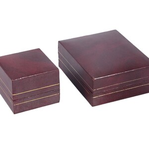 May include: Two burgundy-colored gift boxes with gold trim. The larger box is on the right and the smaller box is on the left.