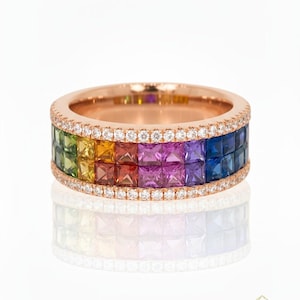 May include: Rose gold ring featuring a band of square-cut gemstones in a rainbow of colors. The ring is accented with a row of small, clear stones and is set against a white background. The ring is a piece of jewelry.