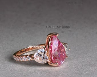 May include: A rose gold ring with a pear-shaped pink gemstone and two smaller pear-shaped clear gemstones. The ring has a band with small clear gemstones.
