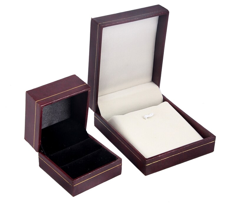 May include: Two burgundy jewelry boxes with gold trim. The larger box is open and has a white velvet interior. The smaller box is closed and has a black velvet interior.