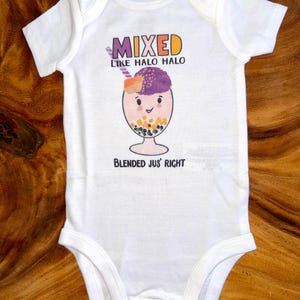May include: White baby onesie with a colorful graphic of a Halo Halo dessert. The text on the onesie reads "MIXED LIKE HALO HALO" above the illustration and "BLENDED JUS' RIGHT" below.
