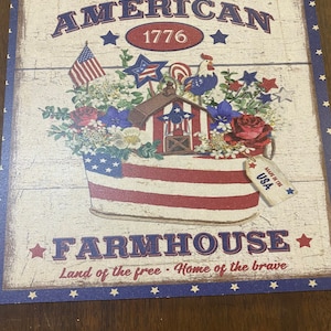 May include: Wooden sign with an American farmhouse theme. The sign features the words "AMERICAN" and "FARMHOUSE" with the year "1776". The design includes an American flag, flowers, and a rooster. The sign is decorated with red, white, and blue colors.