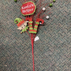 Op de afbeelding: Rode, groene en witte glitter kerstspray met een rood bord waarop "Santas Helper" staat en een groen en rood elfje.