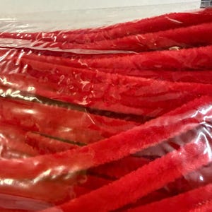 May include: A bundle of bright red pipe cleaners with a soft, fuzzy texture, encased in a clear plastic packaging sleeve. The flexible, cylindrical craft stems are arranged in parallel rows, showing their vibrant color and plush surface material.