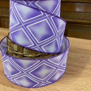May include: Two rolls of purple and white patterned ribbon with a diamond design. The ribbon is on a wooden surface.