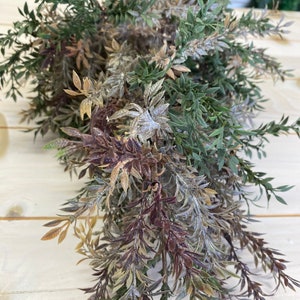 May include: A bunch of artificial greenery with a mix of green, brown, and purple leaves. The leaves are frosted and have a realistic look.