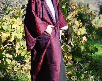 May include: A long, burgundy kimono-style robe with a geometric pattern. The robe is worn over a white shirt and black pants. The sleeves are wide, and the robe has pockets. The background features green foliage and a natural setting.