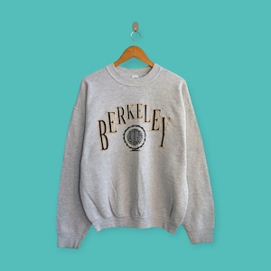 Könnte beinhalten: Ein hellgraues Sweatshirt mit dem Wort "BERKELEY" in schwarzen und goldenen Buchstaben auf der Brust. Ein kreisförmiges Emblem befindet sich unter dem Text. Das Sweatshirt hängt an einem Holzbügel vor einem türkisfarbenen Hintergrund.