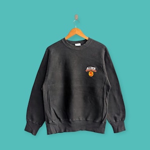 May include: A black crewneck sweatshirt with the text "ALIEN WORKSHOP 5" in red and yellow. The sweatshirt is hanging on a wooden hanger against a teal background. The garment has a vintage, worn look.