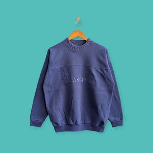 Puede incluir: Sudadera azul marino con la palabra "Australia" bordada en el pecho. La sudadera tiene cuello y puños acanalados. La prenda está colgada de una percha de madera sobre un fondo turquesa.