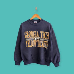 May include: Navy blue sweatshirt featuring the text "GEORGIA TECH YELLOW JACKETS" in yellow and white. The sweatshirt has a crew neck and long sleeves. A graphic of a yellow jacket is between the words.