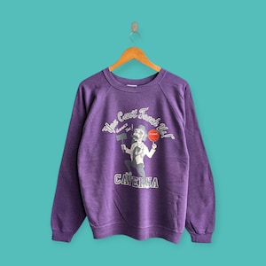 May include: Purple vintage sweatshirt with a graphic of a cartoon man holding a hammer and a ball, with the text "You Can't Touch Us!" and "Caverna". The long-sleeved shirt is hanging on a wooden hanger against a teal background.