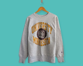 May include: A heather gray sweatshirt with the words "UNIVERSITY MINNESOTA" in gold lettering. The sweatshirt has a circular emblem in the center with a brown background and a crest design. The sweatshirt is hanging on a wooden hanger.