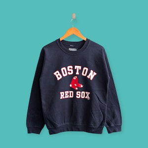 May include: Navy blue sweatshirt featuring the text "BOSTON RED SOX" in white lettering with a red sock graphic. The sweatshirt has a crew neck and long sleeves. The garment is hanging on a wooden hanger against a teal background.