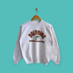 Puede incluir: Sudadera gris con la palabra "Dolphins" en arco marrón y un logotipo de delfín. Debajo está el texto "National Football League". La sudadera está colgada de una percha de madera sobre un fondo turquesa.