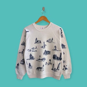 Può includere: Felpa color crema con un motivo ripetuto di illustrazioni di fari blu navy. I fari variano nel design e nelle dimensioni, creando un tema nautico. La felpa ha maniche lunghe e girocollo.