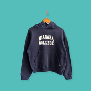 May include: A navy blue hooded sweatshirt with the words "NIAGARA COLLEGE" in white block letters across the chest. The sweatshirt has a front pocket and a drawstring hood. It is hanging on a wooden hanger against a teal background.