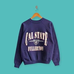 Könnte beinhalten: Ein marineblaues Sweatshirt mit Rundhalsausschnitt und den Worten "CAL STATE FULLERTON" in weißer und roter Schrift. Das Sweatshirt hat einen Vintage-Look und hängt an einem Holzbügel.