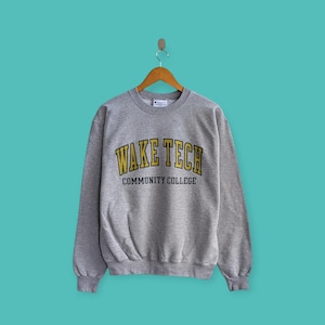 May include: A gray Champion sweatshirt with the text "WAKE TECH COMMUNITY COLLEGE" in yellow and black lettering. The sweatshirt is hanging on a wooden hanger against a teal background.