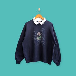 Puede incluir: Sudadera azul marino con cuello blanco. La sudadera presenta un diseño floral bordado con un iris enmarcado. El cuello tiene un pequeño detalle bordado. La sudadera está colgada en una percha de madera.