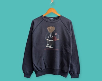 May include: Navy blue sweatshirt featuring a bear wearing a "USA" jersey and holding a basketball. The graphic is centered on the front of the crew neck sweatshirt. The text "POLO BEAR BY RALPH LAUREN" is printed below the bear.