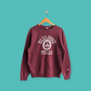 Könnte beinhalten: Ein weinrotes Champion-Sweatshirt mit den Worten "Tarleton Texas" in weißem, bogenförmigem Text. Ein kreisförmiges Emblem mit einem Gebäude befindet sich unter dem Text. Das Sweatshirt hängt an einem Holzbügel vor einem türkisfarbenen Hintergrund.