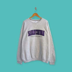 May include: A grey sweatshirt featuring the text "SASKATCHEWAN POLYTECHNIC" in purple and black. The crew neck sweatshirt is suspended on a wooden hanger against a teal backdrop. The Russell Athletic logo is visible.
