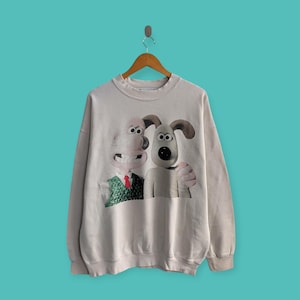 May include: A cream-coloured sweatshirt featuring a graphic of Wallace and Gromit. Wallace is wearing a green waistcoat and red tie. Gromit is embracing Wallace. The sweatshirt is hanging on a wooden hanger against a teal background.