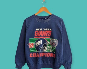 May include: Navy blue sweatshirt featuring the text "NEW YORK GIANTS" in red and white. The design includes a football helmet and the word "CHAMPIONS" in red. A "SUPER BOWL XXV" patch is also visible.