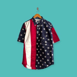 May include: Short-sleeved button-down shirt with a patriotic design. The shirt features a white stripe down the left side, a red stripe on the right, and a navy blue background with white stars. The shirt is hanging on a wooden hanger.