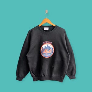 Puede incluir: Sudadera negra con un logotipo vintage de los New York Mets que presenta el nombre del equipo y una pelota de béisbol con el horizonte de la ciudad de Nueva York en el fondo.