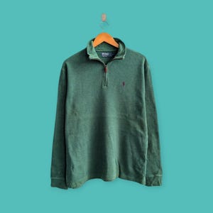 May include: A green long-sleeved sweatshirt with a quarter-zip collar and a small red logo on the chest. The sweatshirt is hanging on a wooden hanger against a teal background. The fabric appears to be a soft, worn material.