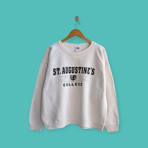 May include: Cream-colored sweatshirt with the text "ST. AUGUSTINE'S COLLEGE" in black. The sweatshirt is hanging on a wooden hanger against a teal background. The text is centered on the chest.