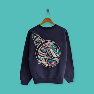 Peut inclure: Sweat-shirt bleu marine avec un motif d'épaulard détaillé dans le dos. Le motif d'épaulard comprend des éléments rouges, bleus, blancs et noirs, créant un visuel saisissant. Le sweat-shirt est suspendu à un cintre en bois sur fond turquoise.