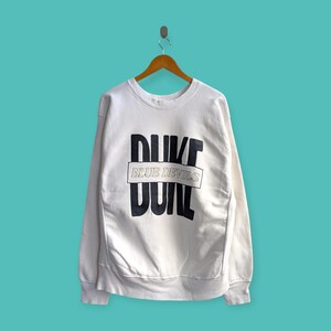 May include: White sweatshirt with the text "DUKE BLUE DEVILS" in black block letters. The sweatshirt is hanging on a wooden hanger against a teal background. The sweatshirt has long sleeves and a crew neck.