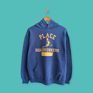 May include: A navy blue hoodie with a kangaroo pocket. The front displays the text "PLACE ROADRUNNERS" in yellow, accompanied by a graphic of a running figure. The sweatshirt is suspended from a wooden hanger.