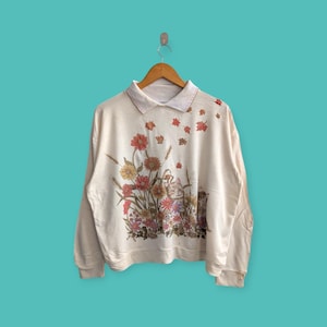 May include: A cream-coloured long-sleeved sweatshirt with a collared neckline. The sweatshirt features a floral print with a variety of flowers and leaves in shades of pink, red, orange, yellow, and green. The print also includes a small image of a cat.