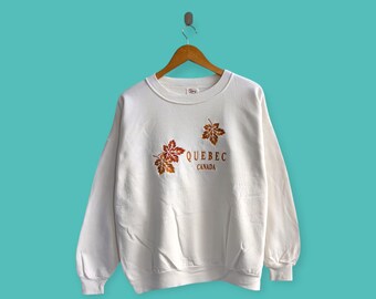 Puede incluir: Sudadera blanca con hojas de arce bordadas en tonos marrón y naranja, con el texto "QUEBEC CANADA". La prenda está colgada de una percha de madera sobre un fondo turquesa.