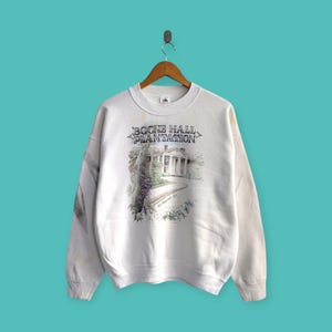 May include: White crew neck sweatshirt with a graphic of Boone Hall Plantation, featuring the text "BOONE HALL PLANTATION" at the top. The graphic includes a detailed illustration of the plantation house and surrounding foliage. The sweatshirt is hanging on a wooden hanger.