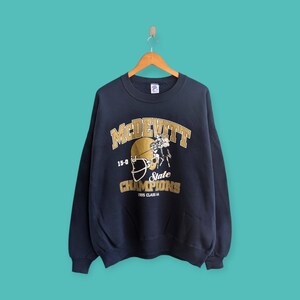 Puede incluir: Sudadera azul marino con letras y gráficos dorados. La parte delantera presenta la palabra "McDevitt" encima de un gráfico de casco de fútbol y las palabras "State Champions 1995 Class A". La sudadera está colgada en una percha de madera.
