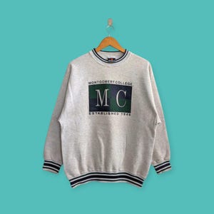 May include: A light gray sweatshirt with the text "MONTGOMERY COLLEGE" above a green and navy blue rectangle with the letters "MC" and the text "ESTABLISHED 1946". The sweatshirt has navy blue and white striped trim.
