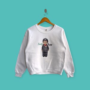 Op de afbeelding: Witte crewneck sweatshirt met een afbeelding van een cartoonjongen in een blauwe overall en een rode bandana. De tekst "Buddy Lee" is op de sweatshirt gedrukt.
